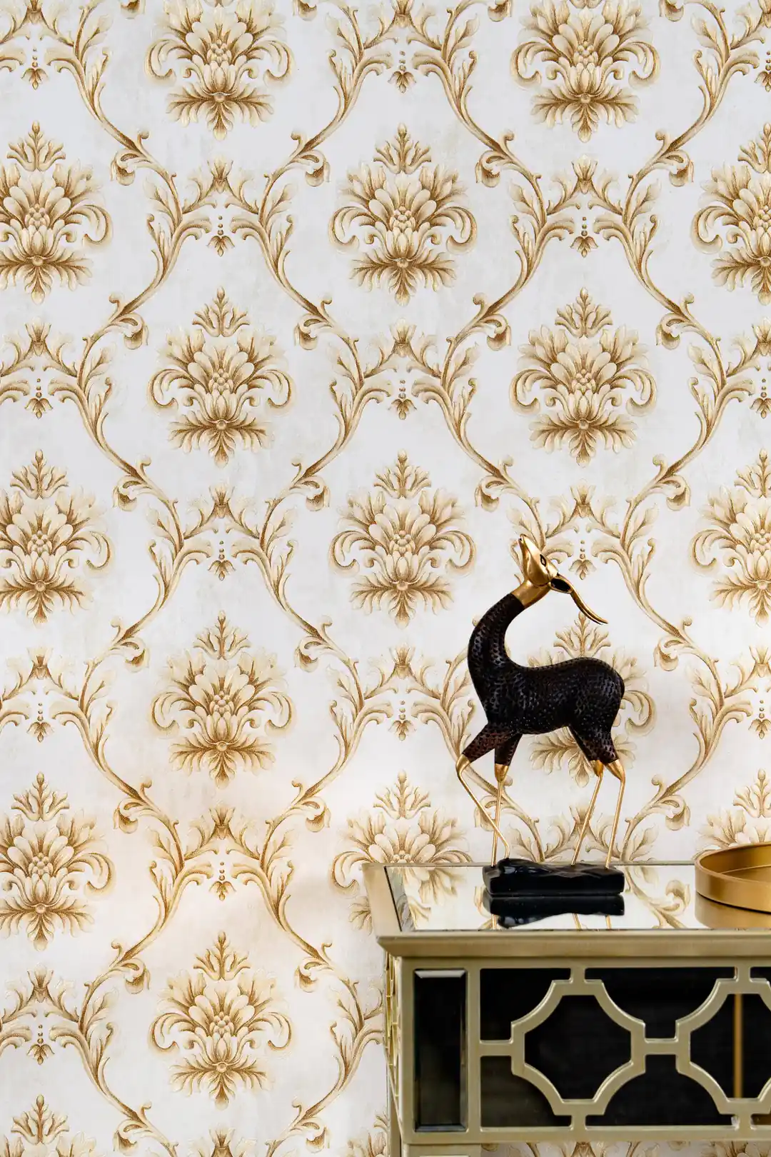 Installed gold baroque wallpaper with ornate damask pattern behind a glass console table and decorative deer figurine