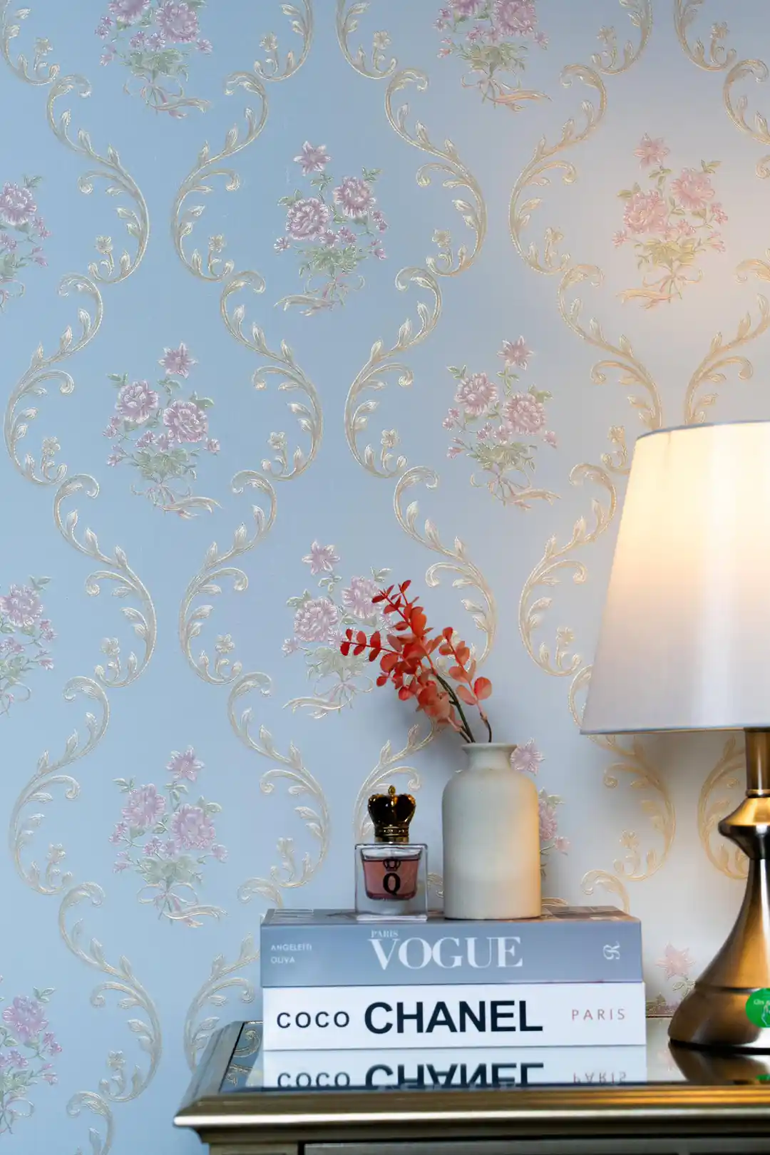 Elegant room vignette with pink floral damask wallpaper behind bedside table with lamp and Vogue books