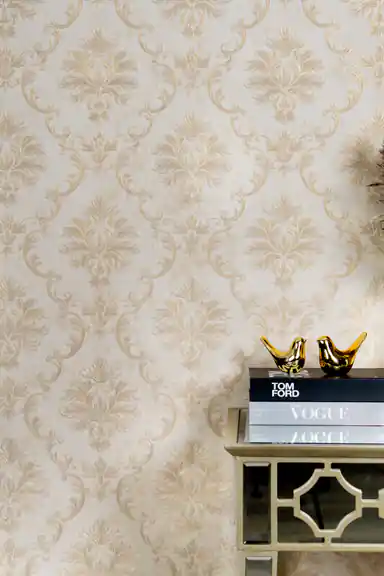 Close-up of embossed golden damask acanthus motif on cream wallpaper with fashion books and golden bird figurines on mirrored shelf
