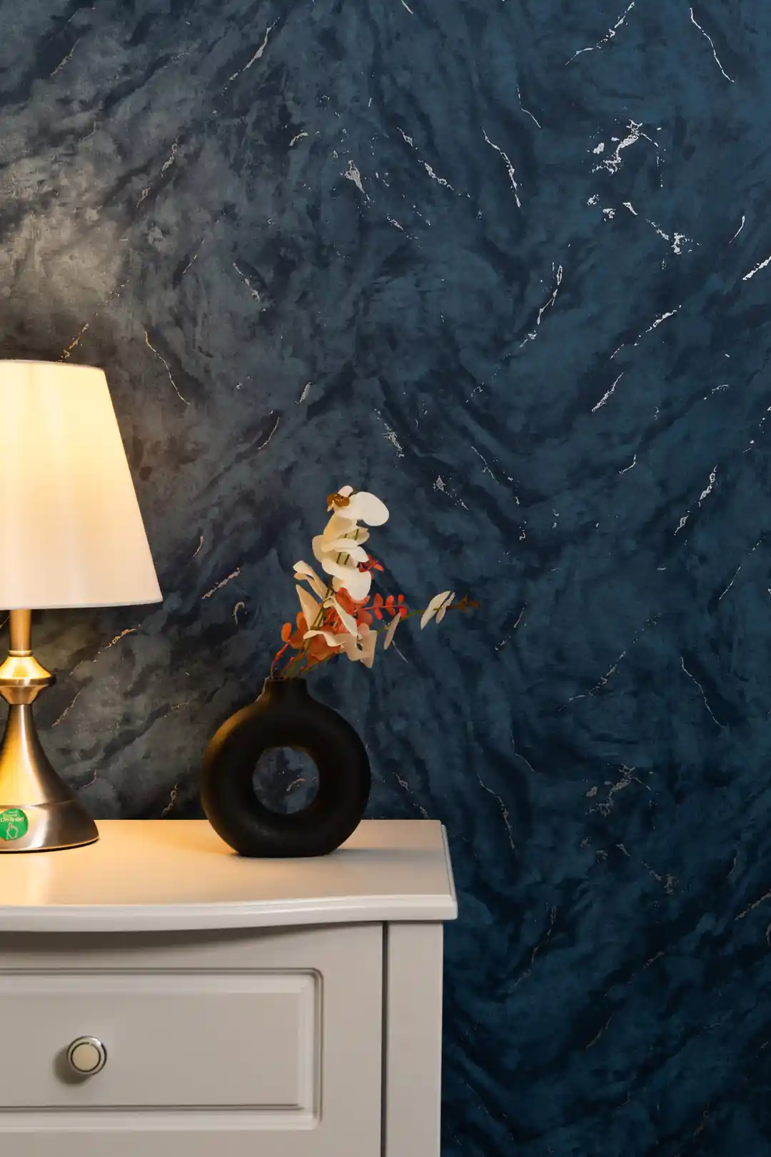 Dark blue marble wallpaper behind bedside table with lamp and decorative vase in cozy setting