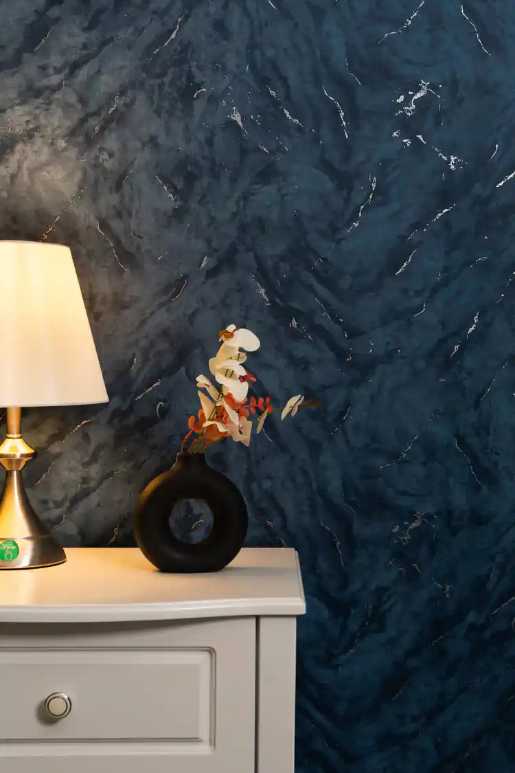 Dark blue marble wallpaper behind bedside table with lamp and decorative vase in cozy setting