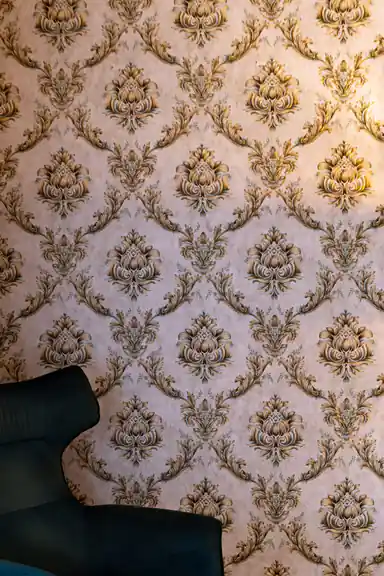 Gold damask wallpaper on cream base behind velvet green armchair with warm lamp lighting in living room