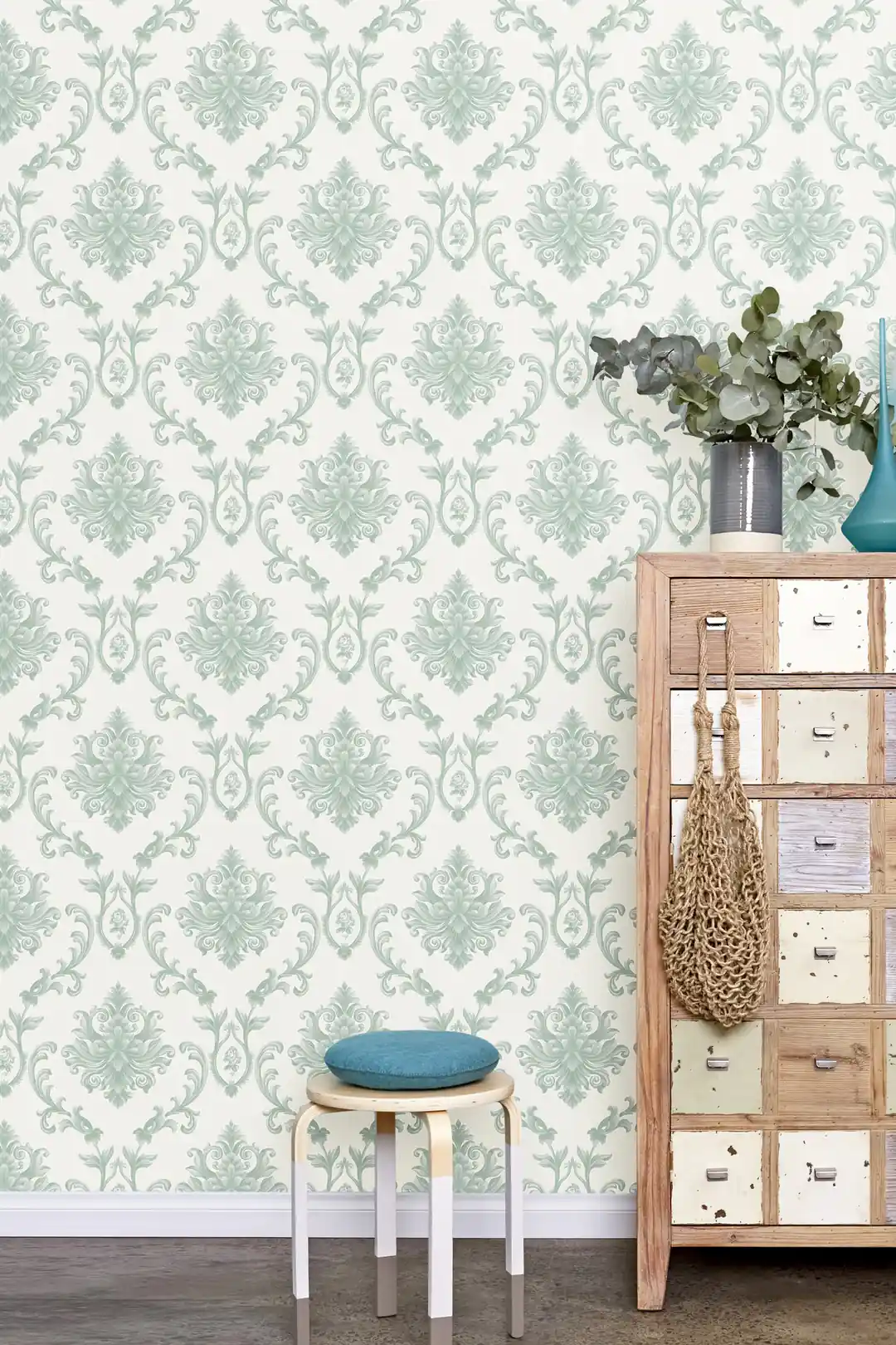 Room scene with sage green damask wallpaper behind a wooden drawer unit, teal stool, and eucalyptus vase