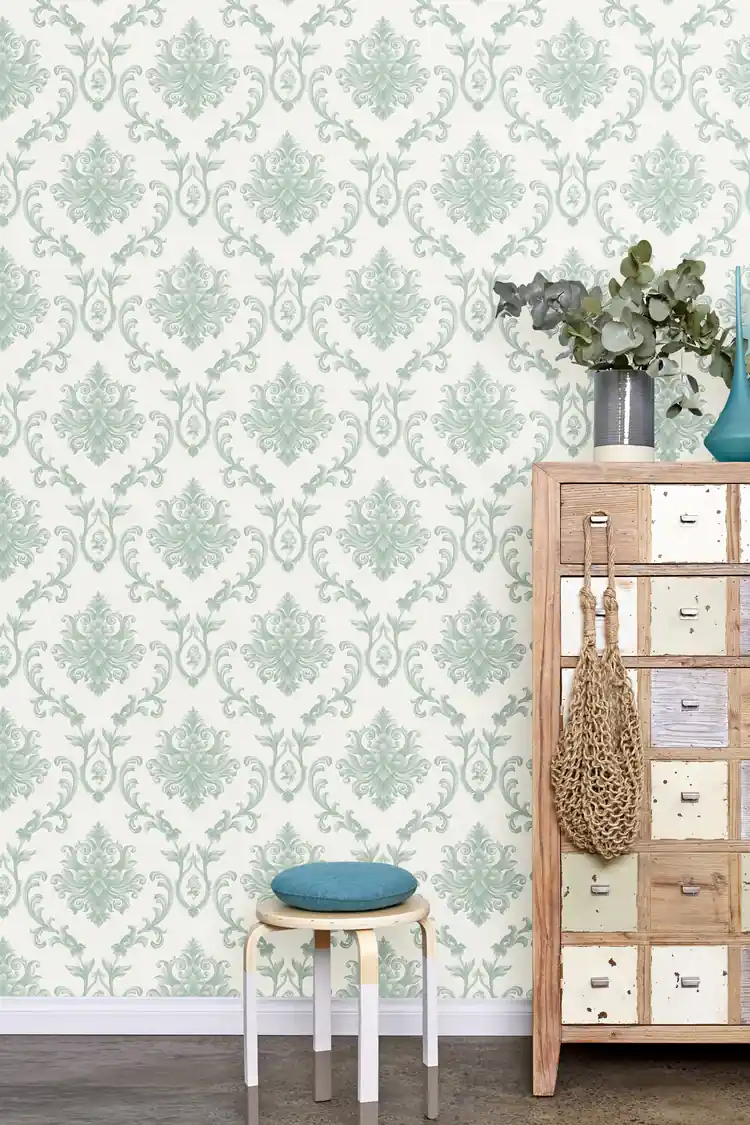 Room scene with sage green damask wallpaper behind a wooden drawer unit, teal stool, and eucalyptus vase