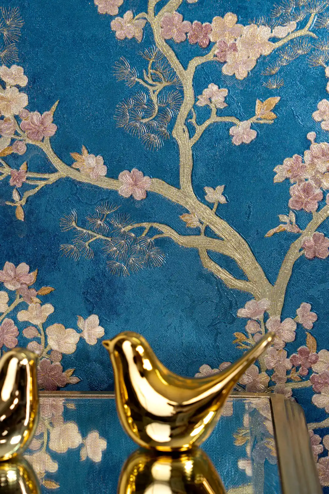 Close-up of golden branches and pink blossoms with a gold bird figurine on blue wallpaper background