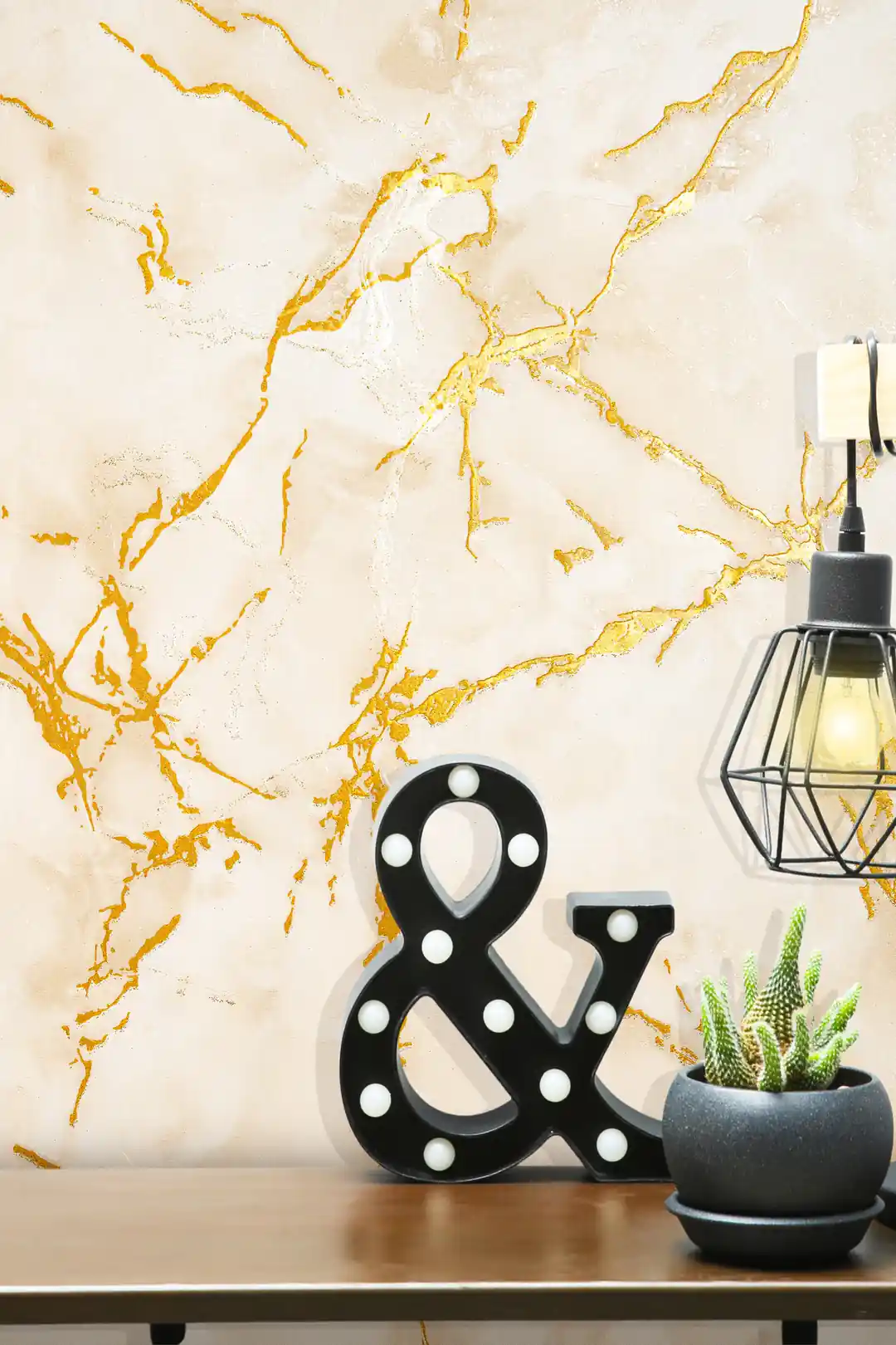 Beige marble wallpaper with golden veining behind a black ampersand marquee light, cactus plant, and cage lamp on a wooden shelf