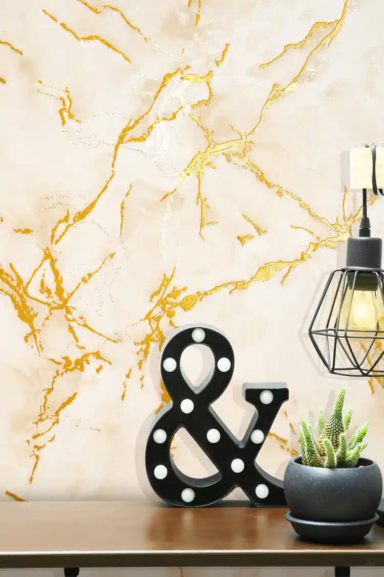 Beige marble wallpaper with golden veining behind a black ampersand marquee light, cactus plant, and cage lamp on a wooden shelf