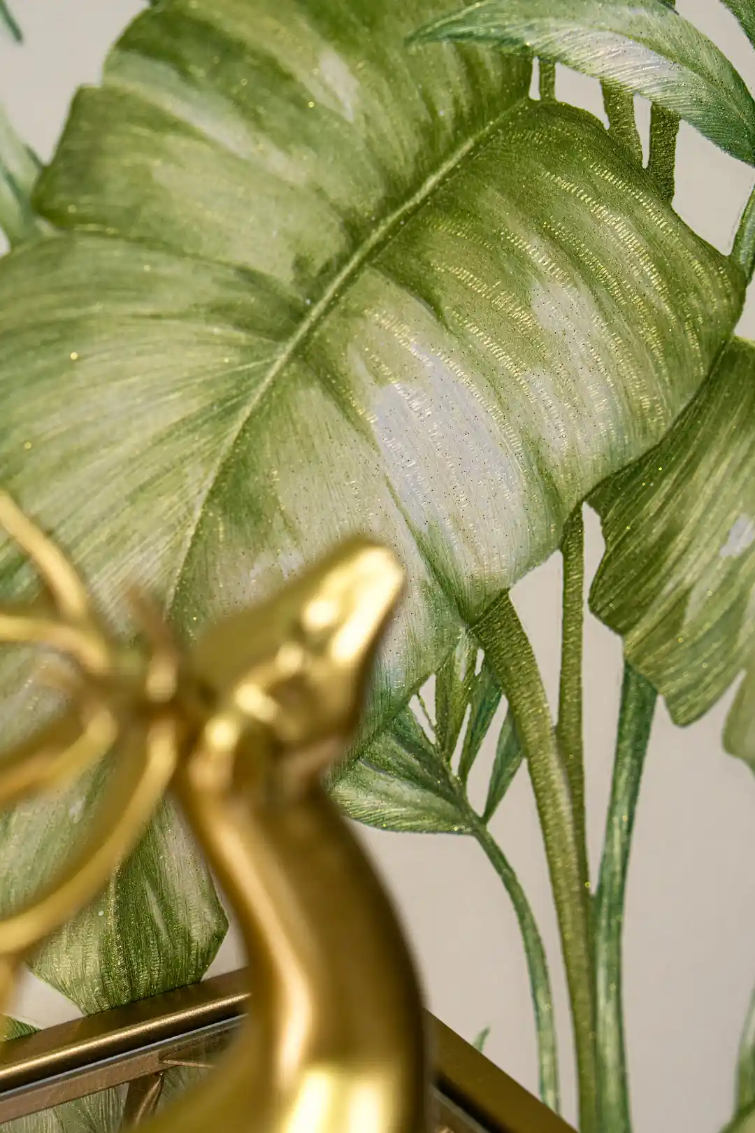 Close-up of green monstera leaf with gold shimmer on cream wallpaper behind a gold abstract figurine