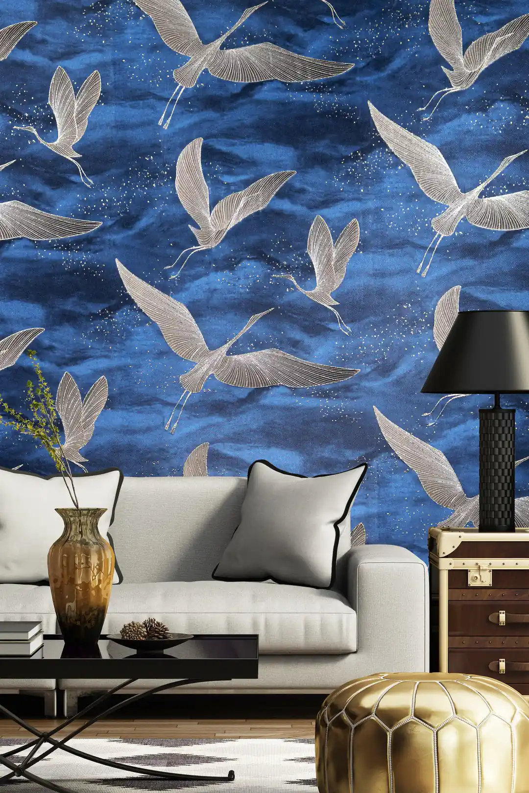 Living room with deep blue crane wallpaper behind a grey sofa with gold pouf, black lamp, and vintage trunk