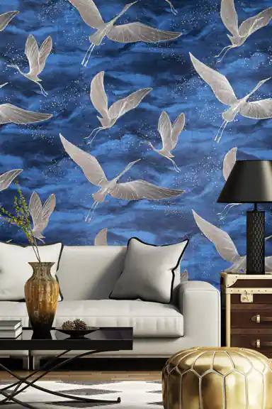 Living room with deep blue crane wallpaper behind a grey sofa with gold pouf, black lamp, and vintage trunk